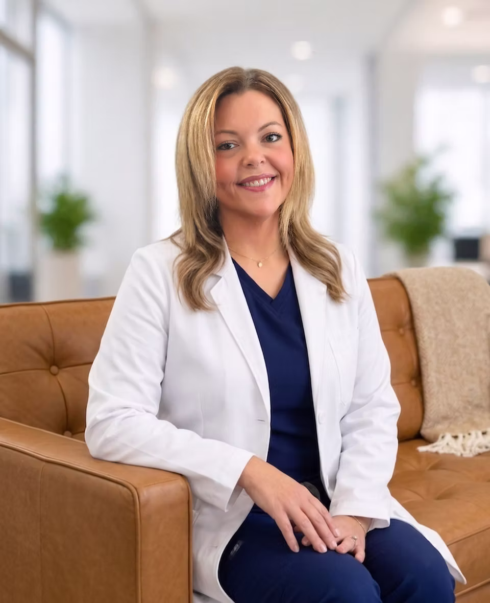 Megan Winters in a white coat sits on a tan couch in a bright office, with blurred plants and windows behind her.